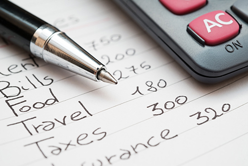 A close-up of a pen on top of a list which includes bills, food, travel and taxes and numbers beside each.