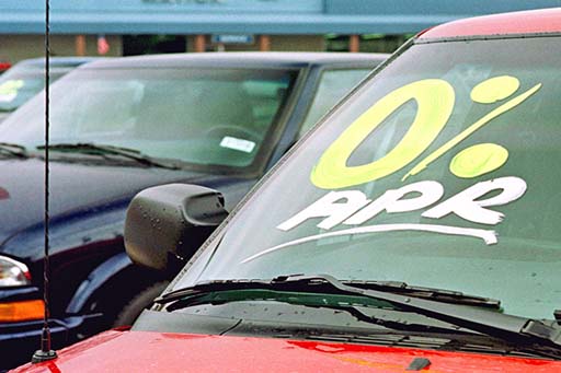 The image is a photo of a car showroom with three cars in shot. The car on the right-hand side has ‘0% APR’ written in large print and underlined on the windscreen.