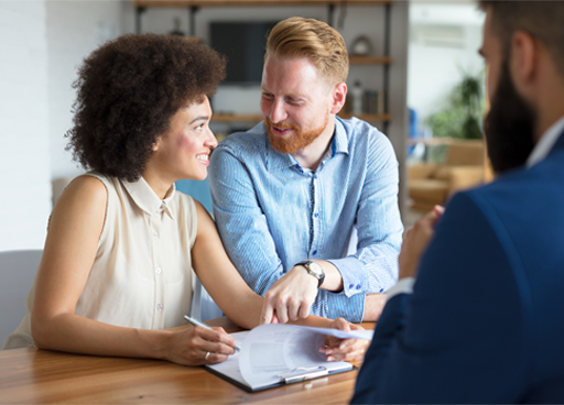 The image is a photograph of a young couple completing paperwork (relating to a financial transaction) with an adviser, who is sitting on the other side of the desk.