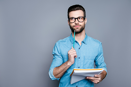 The image is a photo of a young man holding a large note book and a pencil whilst deep in thought.