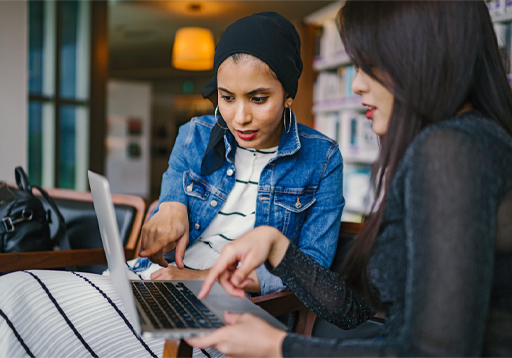 The image is a photograph of two women with a laptop open in front of them. They are both looking at the screen intently with one woman pointing at the screen. Behind them is a bookshelf which is full of books.
