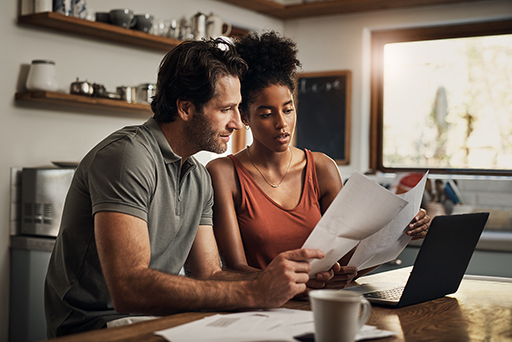 The image is a photo of a young couple studying paperwork, with a laptop computer open on a kitchen ‘island’.
