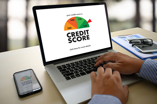 The image shows an open lap top with the words ‘Credit Score’ on the screen with an image of a semi-circular indicator (from low credit score on the left-hand side to a high credit score on the right-hand side). The person using the laptop has a high credit score. Elsewhere on the desk are a mobile ‘phone (with the same image on the screen as the laptop) , some documents on a clipboard, a notebook and spectacles.