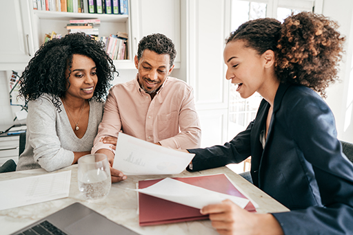 The image is a photo of a young couple in their home in discussions with their (female) financial adviser. The couple are reviewing financial planning documents provided by the adviser.