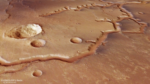 This figure shows the surface of Mars viewed at an angle from above. The surface is coloured shades of red-brown. A winding branching pattern goes across the image from bottom left to top right. Four circular features are visible, of different sizes.