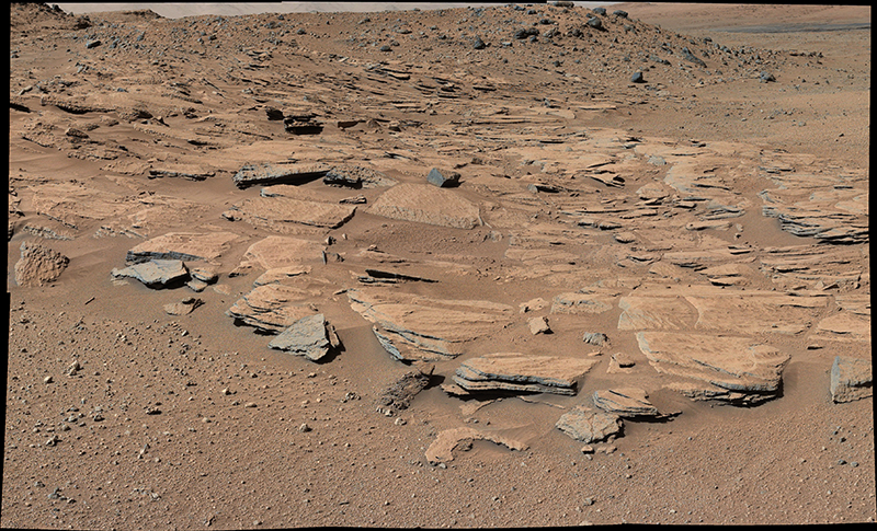This figure is a photograph of the martian surface taken by the Curiosity rover. In the foreground is an area of fine grained martian dust with some larger pebbles. Behind this is a rock outcrop covered by a thin layer of dust. The dust is a red-brown colour, and the rock outcrop is green-orange. The rock has horizontal, parallel beds.