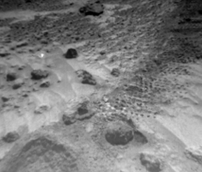 This figure is a photograph taken by the Pathfinder lander of the martian surface. It is a greyscale image. It shows fine grained dust that contains small rounded pebbles and larger rounded boulders.