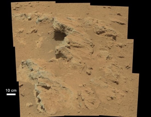 This figure is a photograph of the martian surface taken by the Curiosity rover. It shows a rock outcrop covered by a thin layer of dust. The dust is a red-brown colour, and the rock outcrop is green-orange.