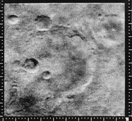 This figure shows a photograph taken from orbit of the martian surface. It is in greyscale and is heavily pixelated. Several circular features are evident, which are craters.