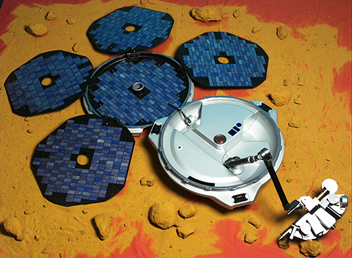 This figure is a photograph of the beagle 2 lander. It shows a circular silver disk with an arm protruding outwards to the right. On the end of the arm is a metallic, semi-circular shaped box housing scientific instruments. On the opposite side of the silver disk to the arm a blue circular disk is attached. Four roughly circular, blue disks are attached to its circumference. These five circular disks are solar panels.
