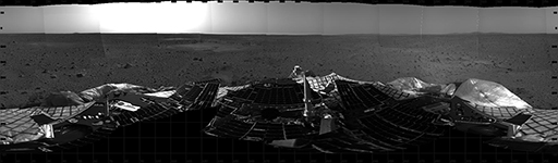 This figure is a panoramic photograph taken by the Spirit rover of the martian surface. It is in greyscale. In the foreground is part of the rover’s landing platform. In the background is a dark grey surface with fine grained material and some boulders evident. The horizon is flat and sky is a pale grey colour.