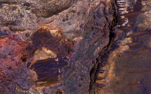 This figure is a photograph taken from Mars orbit. The main feature is a ridge extending north to south on the right hand side of the image. To the right of this ridge, dark brown horizontal lines are shown, surrounded by purple and brown material. To the left of this ridge are complex shapes with different textures and colours.
