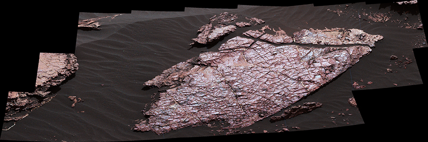 This figure is a photograph taken by the Curiosity rover looking down on the top of a rock outcrop on the martian surface. The rock outcrop is surrounded by fine grained, dark grey soil. The surface of the rock appears to be cracked.