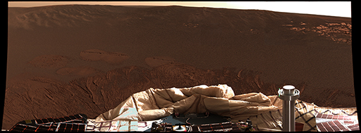 This figure is a photograph taken by the Opportunity rover of the martian surface. It is in shades of orange-brown. In the foreground is part of the rover’s deflated airbags. In the background is a dark brown surface with fine grained material and some boulders evident. In the distance, rock outcrops can be seen. The horizon slopes slightly to the right hand side and the sky is pale orange.
