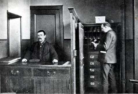 This black and white photograph depicts two men in an office. One of them sits behind a reception desk on the left, and the other stands beside a filing cabinet on the right. There is a wooden partition between them.