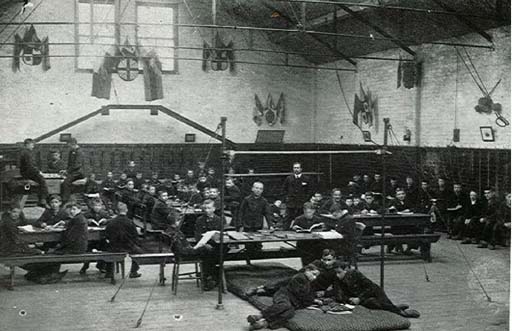 In the foreground of this black and white photograph, three boys lie on a gym mat underneath a high-bar frame, studying something in front of them. Several long desks are arranged behind them, with boys sitting on benches at either side. A schoolmaster stands to the right of centre, looking towards the camera.