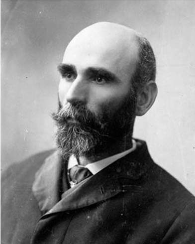 This is a black and white head and shoulders photograph of a bearded man with a receding hairline. He faces to the left, with a serious expression and a steady gaze. He wears an overcoat buttoned to the collar, over a white shirt and tie.