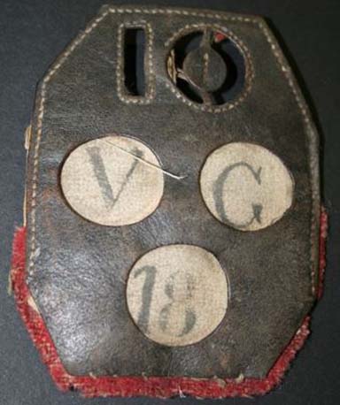 This is a colour photograph of a tooled leather badge. The number 10 is cut into the leather at the top, with the letters VG and the number 18 printed in white circles beneath it. The remains of a red fabric border adhere to the bottom of the badge.