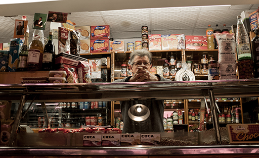 A man stands behind a counter on a market stall. There are a range of drinks and snacks are available.