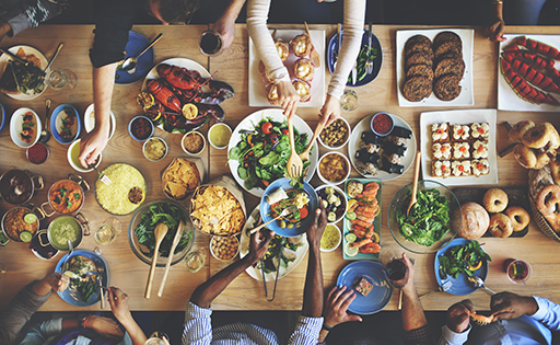 A top down shot of a table containing a broad range of foods from around the world. People are reaching in from all angles to share the food.