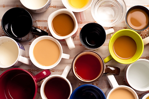 A top-down shot of a table covered in mugs of all colours. The mugs are all filled with different types of liquid (presumably different types of tea and coffee)