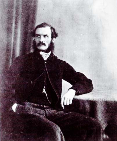 This black and white photographic portrait features a man seated with one elbow propped on a table. He faces to the right, with his head turned to the left. He has dark hair, a moustache and full sideburns, and wears a velvet jacket over a buttoned waistcoat. A metal implement is fastened to one of his waistcoat buttons.