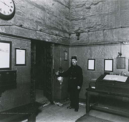 This is a black and white photograph of a uniformed man standing beside an open doorway. He is in a high-ceilinged room, whose walls are hung with framed documents. What looks like a visitor book lies open on a desk to the right, and there is a clock on the wall at upper left.