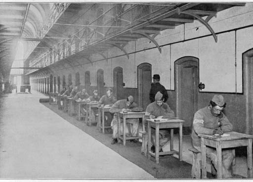 This black and white photograph depicts the interior of an extensive cell block, receding from the right foreground. Outside each cell door sits a man at a wooden desk, their heads bent forward as they write. A warder walks alongside them, supervising the activity.