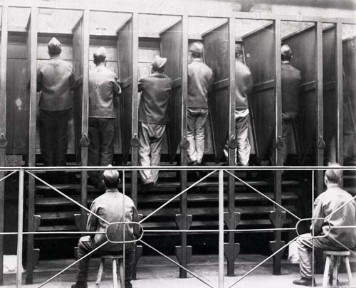 In this black and white photograph, six men in prison clothing are mounted on a treadmill, with their backs to the viewer. They are separated from each other by wooden partitions. Two other prisoners sit on wooden stools in the foreground, watching the treadmill turn.