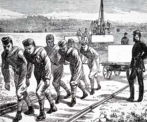 This is a drawing of six convicts hauling a handcart loaded with a large stone block along metal rails. The men trudge towards the left foreground, leaning forward into the weight of the task. Two more carts are loaded by crane in the background, while a supervisor looks on from the right.
