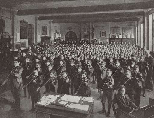 This is a black and white photograph of a large school hall packed with standing boys, who are supervised by teachers at the sides of the room. A boy sits at a piano in the right foreground, with three rows of schoolfellows behind him forming an orchestra of stringed and wind instruments.