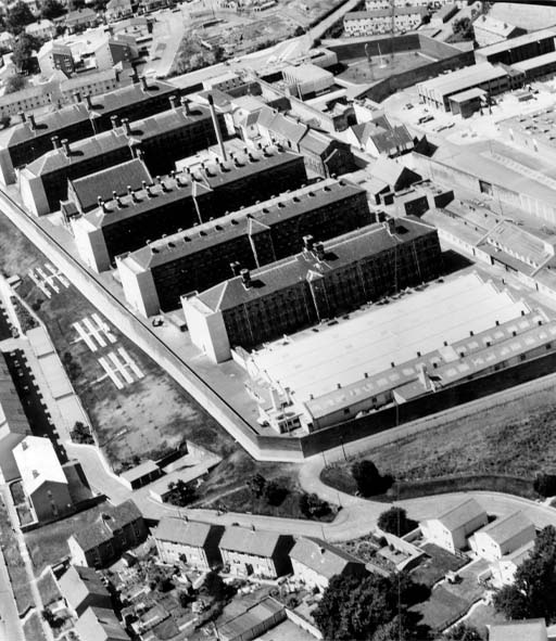 This black and white photograph provides an overhead view of a walled prison complex. It consists of five separate four-storey blocks of identical size, with a chapel between two of them at upper left. There are what look like workshops at lower right, and an open yard at upper right.