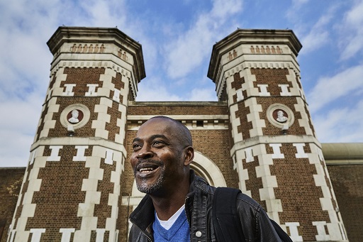 This is a colour photographic portrait of a smiling man, framed against the elaborately designed frontage of a prison entrance. He faces to the left, and wears a grey denim jacket over a blue t-shirt. The shot is taken from below, so that we see him in a head and shoulders view, with the prison towers outlined against a blue sky.