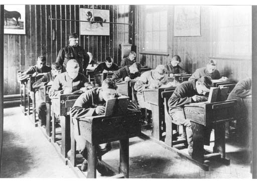 A dozen boys are seated at three lines of desks in this black and white photograph. Their heads are bent forward as they apparently copy text from the books propped open in front of them. They are supervised by a teacher wearing what looks like military uniform.