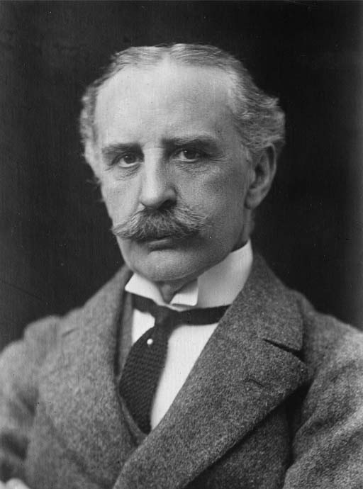 This is a black and white photographic portrait of a man with a bushy moustache. Framed in a head and shoulders view, he wears a heavy wool jacket over a high-collared white shirt, and gazes straight at the camera.