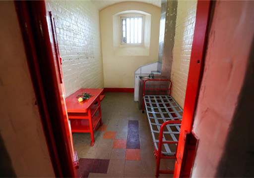 This is a colour photograph of a prison cell, preserved as it was in the past. A metal bunk with a strapped bed-base stands against the right-hand wall, with a red-painted writing desk opposite. The floor of the cell is tiled, and the brick walls painted cream. Light enters the cell through a window at the far end.