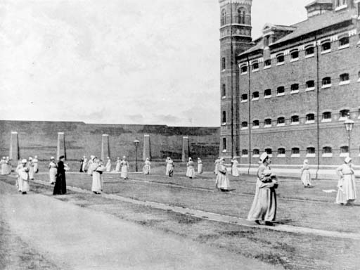 This is a black and white photograph of women walking in a prison yard, bordered by a cell block on the right. Each woman walks at a distance from the next, and some are cradling babies in their arms. They wear loose, light-coloured dresses, with white caps covering their hair.