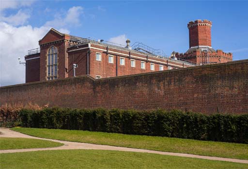 This is a colour photograph of a red-brick prison building, mostly concealed by a high exterior wall. Small windows can be seen at the upper level, with a wide octagonal chimney rising above the building on the right.