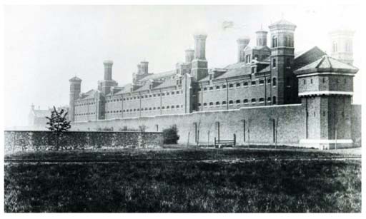 This is a black and white photograph of a prison building, partly concealed behind a high wall. Cell windows line the upper levels of the building, and massive chimneys rise above its roofs. There are observation towers at each end of the prison’s visible façade.
