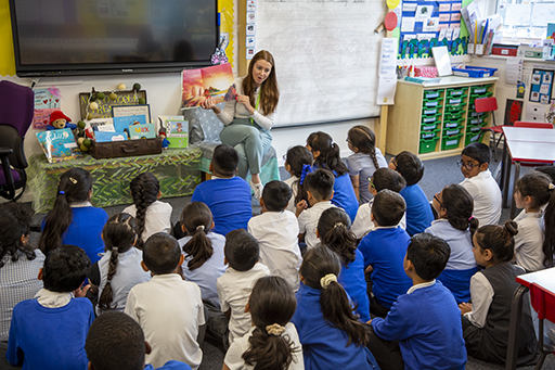 A class of children sat on the floor listening to their teacher reading a book.