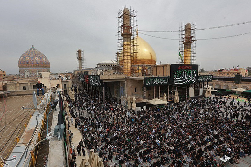 This is a colour photograph of the Al-Askari Mosque, taken in 2017. In front of the mosque there is a large gathering of people. The mosque’s two minarets are covered in scaffolding. Behind them the mosque’s large and shiny golden dome has been repaired. The building is draped with black banners decorated with white text in Arabic.