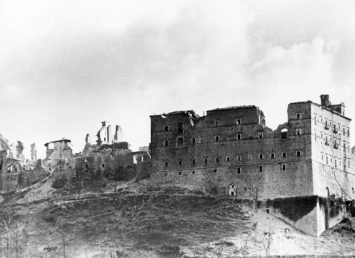 This is a black-and-white photograph taken in nineteen forty-four. It shows Monte Cassino Abbey after the bombing. On the right there is a square building which has several large craters in its roof and walls. To the left there is another connected building which is almost entirely ruined. All that remains are a few walls rising unsupported from the rubble.
