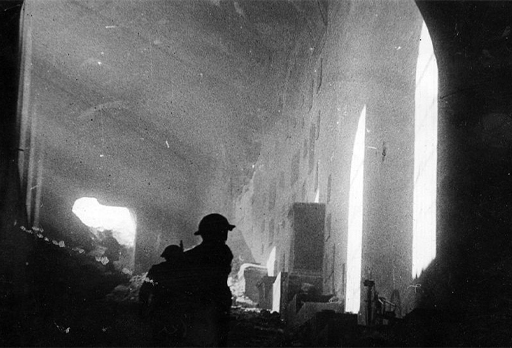 This is a black-and-white photograph taken in nineteen forty-four. It shows the interior of one of the bombed sections of building at Monte Cassino Abbey. Light streams into a long corridor from the left-hand side. In the foreground, a Polish soldier is silhouetted against the rubble behind him.
