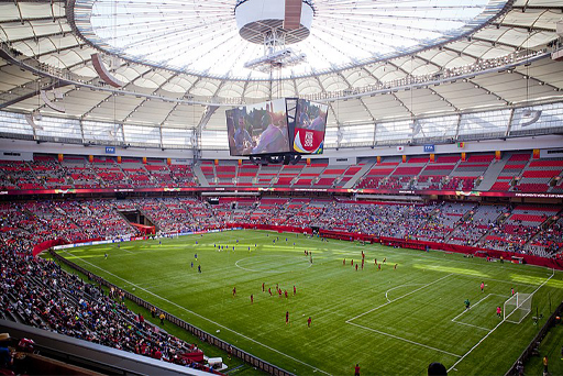 This is a colour photograph of a football stadium. From the perspective of a seat high-up in one of the corners, it shows a large curved section of red seating wrapped around a rectangular grass football pitch. Above the pitch there is a white translucent roof and a large set of screens hanging in the centre. On the pitch, two teams of women footballers are warming up.