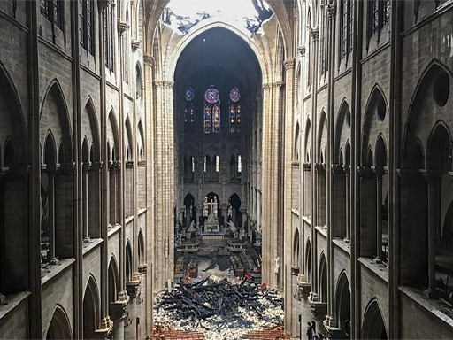 This is a colour photograph taken from inside Notre-Dame Cathedral, near the entrance. It looks east and upward, showing two large holes in the roof of the high vaulted ceiling. Sunlight pours through these holes, illuminating the floor of the church, which is covered with wooden pews and rubble. Along the side of the hall are three floors of ornate stone arches and three stained-glass windows can be seen near the top of the far east wall.