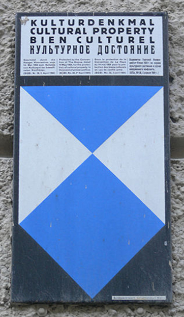 This is a colour photo of the Blue Shield emblem. It shows the emblem attached to a stone wall and labelled in German. The logo takes the shape of a pointed shield divided by two intersecting diagonal lines, which create three triangles at the top of the shield and a single square at the bottom. The square and top triangle are light blue, while the two side triangles are white.