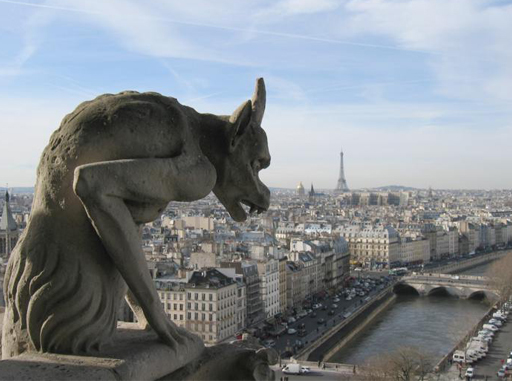 This is a colour photograph of one of Notre-Dame’s gargoyles. It is taken from the roof, looking out over the Parisian skyline, with the Eiffel Tower visible in the distance. The gargoyle is a hunched-over creature, with horns, fangs and slender arms.