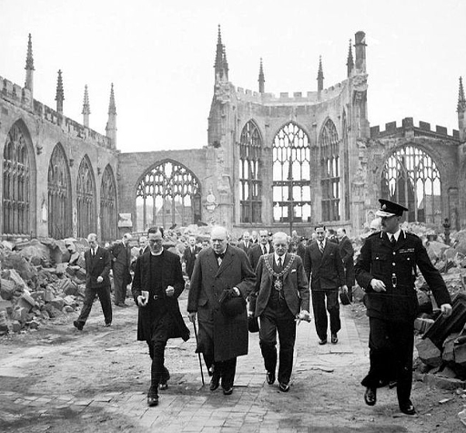 This is a black-and-white photograph taken in nineteen forty. It shows the ruins of Coventry Cathedral from the inside. The entire roof is missing and all of its windows have been broken. Piles of rubble have been moved to the sides of the space by the walls. In the centre, the Prime Minister, Winston Churchill, is walking towards the camera, guided by a policeman and a vicar, with several other suit-wearing men following behind him.
