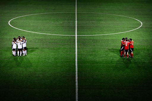 Image of two football teams in two huddles on each side of the halfway line of a football pitch