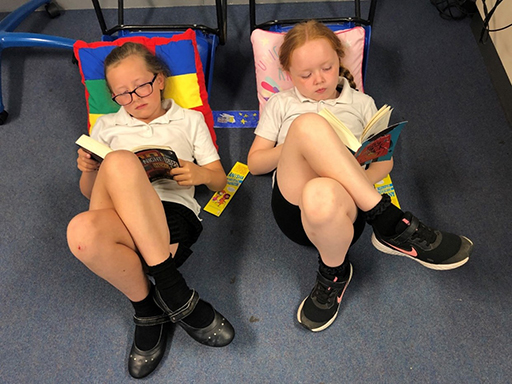 Two girls in school uniform lounging against cushions eading a book each.